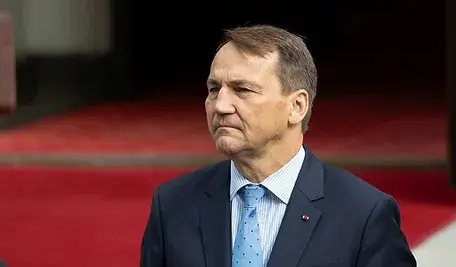 Radosław Sikorski, minister spraw zagranicznych Polski, w granatowym garniturze i błękitnym krawacie, stoi na tle czerwonego dywanu przed wejściem do budynku rządowego.