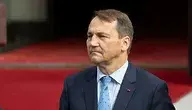 Radosław Sikorski, minister spraw zagranicznych Polski, w granatowym garniturze i błękitnym krawacie, stoi na tle czerwonego dywanu przed wejściem do budynku rządowego.