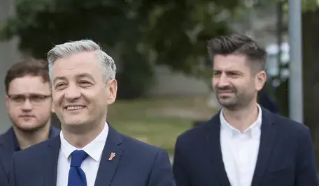 Robert Biedroń i Krzysztof Śmiszek podczas wydarzenia politycznego – liderzy Nowej Lewicy w formalnych garniturach, uśmiechnięci na tle zieleni.