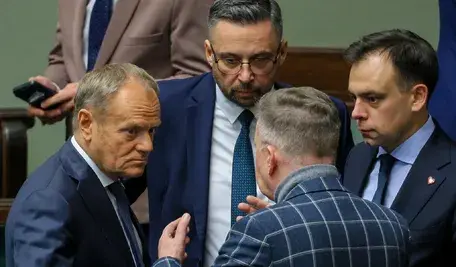 Donald Tusk w otoczeniu swoich ministrów. Ubrany w kraciastą marynarkę Marcin Kierwiński coś mu tłumaczy silnie gestykulując, Tusk, zdaje się być znudzony, a Andrzej Domański wpatrzony jest w Tuska jak w obrazek
