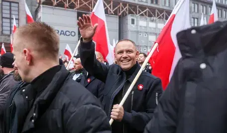 Uczestnicy marszu z biało-czerwonymi flagami w centrum Warszawy, prezydent Karol Nawrocki na pierwszym planie uśmiecha się i macha do tłumu.