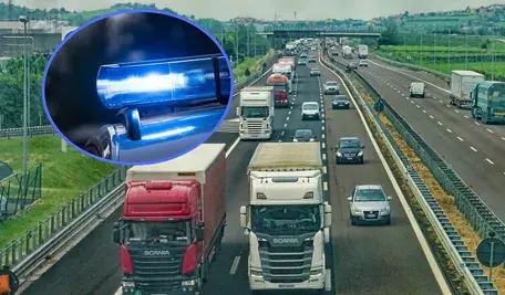 Radiowóz z włączonym niebieskim sygnałem świetlnym na tle ruchliwej autostrady pełnej samochodów ciężarowych i osobowych.