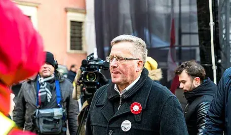 Były prezydent Bronisław Komorowski w płaszczu i okularach stoi wśród uczestników wydarzenia publicznego, w tle widać kamery i reporterów.