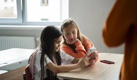 Dwie dziewczynki z telefonem w szkole. Od roku 2025/26 uczniowie portugalskich gimnazjów i liceów nie będą mogli korzystać ze smartfonów w szkołach. Minister edukacji zapowiada wsparcie dla cyfrowej edukacji opartej na sztucznej inteligencji i alternatywne formy spędzania czasu podczas przerw.