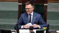 Szymon Hołownia w garniturze siedzący na fotelu marszałka Sejmu podczas obrad, uśmiechający się i trzymający kartkę.