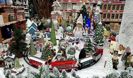 Świąteczna makieta przedstawiająca zimowe miasteczko z dekoracjami bożonarodzeniowymi: miniaturowe domki, ośnieżone drzewka, figurki postaci, kolorowe światełka oraz jeżdżąca kolejka wśród śnieżnej scenerii, ustawiona na sklepowej ekspozycji.