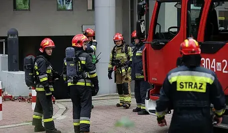 Na zdjęciu widać wóz strażacki na tle budynku, wokół którego stoi sześciu umundurowanych strażaków biorących udział w akcji gaśniczej.