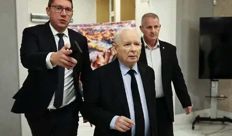 Jarosław Kaczyński w garniturze idzie korytarzem w towarzystwie dwóch innych mężczyzn w formalnych strojach. Jeden z nich wskazuje coś ręką przed siebie.