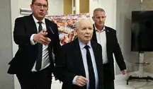 Jarosław Kaczyński w garniturze idzie korytarzem w towarzystwie dwóch innych mężczyzn w formalnych strojach. Jeden z nich wskazuje coś ręką przed siebie.