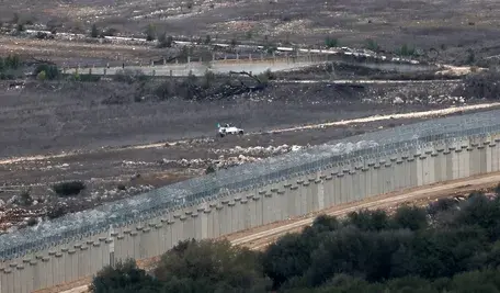 Patrol UNIFIL przy granicy libańsko-izraelskiej obok wysokiego muru bezpieczeństwa, widok na zdemilitaryzowaną strefę i pojazd ONZ na tle pustynnego krajobrazu.”