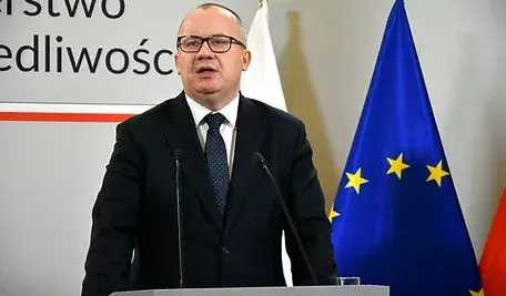 Adam Bodnar przemawia przy podium podczas konferencji prasowej; za nim widoczne są flagi oraz tablica z napisem ministerialnym.