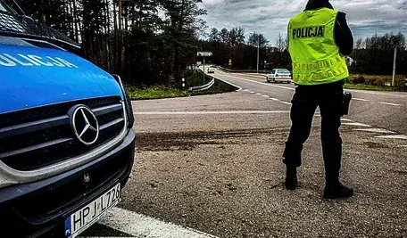 Policjant w żółtej kamizelce na skrzyżowaniu dróg podczas kontroli, obok radiowóz policyjny