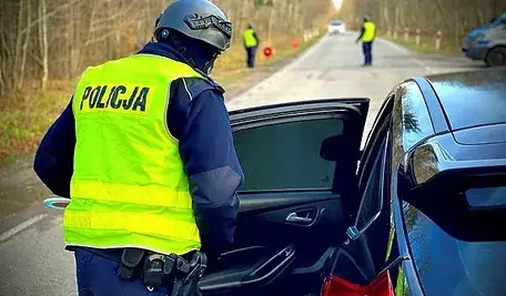 Polski policjant w żółtej kamizelce kontroluje samochód na leśnej drodze, a w tle stoją inni funkcjonariusze.