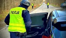 Polski policjant w żółtej kamizelce kontroluje samochód na leśnej drodze, a w tle stoją inni funkcjonariusze.