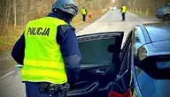 Polski policjant w żółtej kamizelce kontroluje samochód na leśnej drodze, a w tle stoją inni funkcjonariusze.