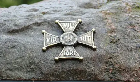 Metalowa płaskorzeźba Orderu Virtuti Militari umieszczona na kamieniu, przedstawiająca krzyż z polskim orłem pośrodku.