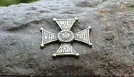 Metalowa płaskorzeźba Orderu Virtuti Militari umieszczona na kamieniu, przedstawiająca krzyż z polskim orłem pośrodku.