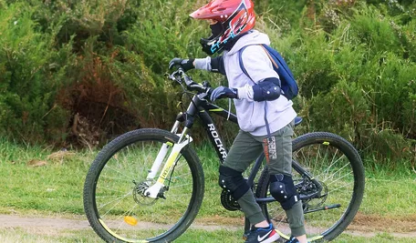 Dziecko w kasku i ochraniaczach prowadzące rower w terenie zielonym.