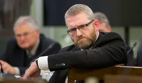 Grzegorze Braun siedzi przy stole, pochylony do przodu, z poważnym wyrazem twarzy.