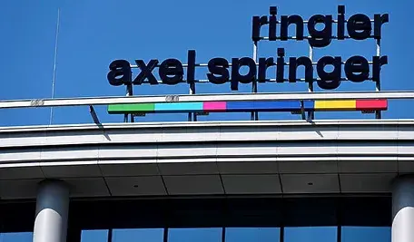 Logo firmy Ringier Axel Springer umieszczone na dachu nowoczesnego budynku, na tle jasnego, bezchmurnego nieba.