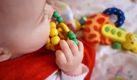 Niemowlę trzyma kolorową zabawkę z drewnianymi koralikami; w tle widać inne dziecięce zabawki.