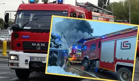 Wozy strażackie podczas akcji gaśniczej; w tle gęsty, ciemny dym unoszący się nad płonącą halą. Na pierwszym planie czerwony wóz straży pożarnej jadący ulicą, a na drugim – strażacy i sprzęt gaśniczy pracujący przy dużym pożarze.