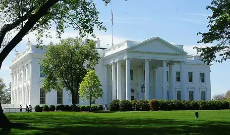 Widok na Biały Dom w Waszyngtonie, otoczony zielonym trawnikiem i drzewami w słoneczny dzień.