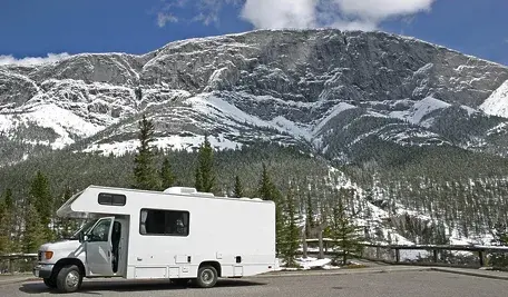 Biały kamper zaparkowany na górskim parkingu, na tle masywnego, ośnieżonego pasma górskiego i lasu iglastego pod jasnym, częściowo zachmurzonym niebem.
