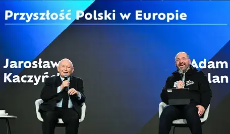 Jarosław Kaczyński i Adam Bielan siedzą na scenie podczas publicznej dyskusji. Obaj trzymają mikrofony i uśmiechają się, a za nimi widoczna jest duża plansza z napisem dotyczącym przyszłości Polski w Europie.