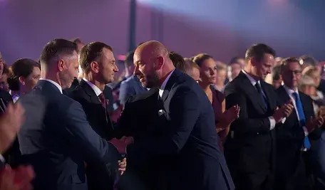 Politycy podczas oficjalnego wydarzenia. Dwaj mężczyźni na pierwszym planie podają sobie ręce, a wokół nich stoją inni uczestnicy, bijąc brawo. Scena odbywa się w sali o uroczystym oświetleniu.