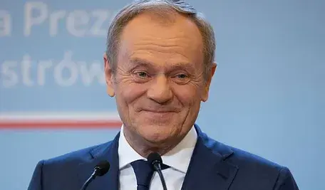 Uśmiechający się Donald Tusk w garniturze stoi przed mikrofonami na tle niebieskiej tablicy z napisem.