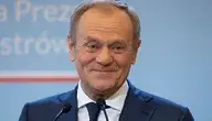 Uśmiechający się Donald Tusk w garniturze stoi przed mikrofonami na tle niebieskiej tablicy z napisem.
