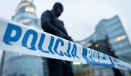 Taśma policyjna i uzbrojony funkcjonariusz w kominiarce zabezpieczający teren przed nowoczesnym budynkiem.