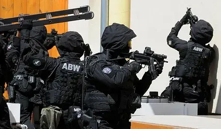 Uzbrojeni funkcjonariusze Agencji Bezpieczeństwa Wewnętrznego w czarnych mundurach prowadzą operację, celując bronią i korzystając z drabin przy wejściu do budynku.