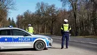 Dwóch policjantów drogówki stoi przy radiowozie zaparkowanym przy poboczu, obok dwupasmowej drogi otoczonej drzewami.