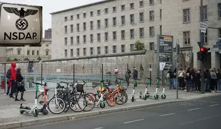 Fragment muru berlińskiego i ludzie na ulicach. W tle orzeł III Rzeszy z podpisem NSDAP.