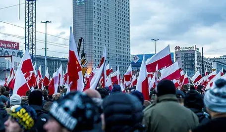 Tłum ludzi z biało-czerwonymi flagami podczas zgromadzenia w centrum miasta; w tle wysokie budynki i reklamy.