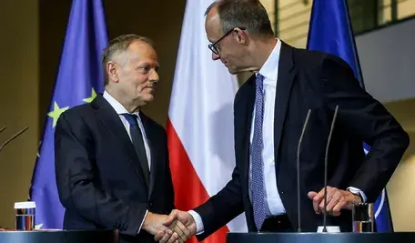 Dwóch ubranych w garnitury polityków stoi obok siebie na tle flag Polski i Unii Europejskiej, podając sobie ręce podczas oficjalnej konferencji prasowej.