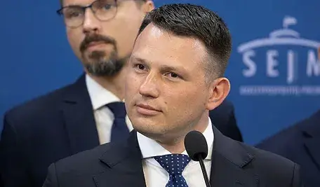 Sławomir Mentzen w garniturze i krawacie stoi przy mównicy na tle niebieskiej ścianki z logo Sejmu; za nim widoczny jest drugi mężczyzna w okularach.