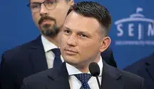 Sławomir Mentzen w garniturze i krawacie stoi przy mównicy na tle niebieskiej ścianki z logo Sejmu; za nim widoczny jest drugi mężczyzna w okularach.