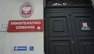 Wejście do budynku Ministerstwa Zdrowia z czerwonym szyldem i godłem Polski nad drzwiami.