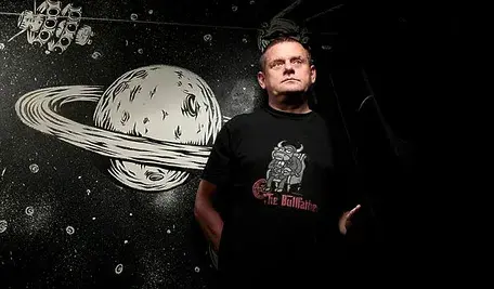 Kazik, wokalista zespołu Kult, stojący na tle czarno-białej, kosmicznej ilustracji przedstawiającej planetę z pierścieniami; ma na sobie czarną koszulkę z grafiką i patrzy lekko w górę.
