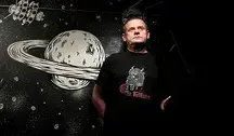 Kazik, wokalista zespołu Kult, stojący na tle czarno-białej, kosmicznej ilustracji przedstawiającej planetę z pierścieniami; ma na sobie czarną koszulkę z grafiką i patrzy lekko w górę.