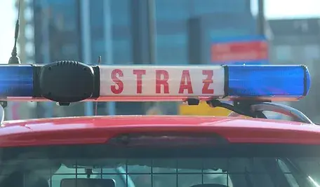 Belka świetlna z napisem ‘STRAŻ’ na dachu samochodu straży pożarnej, widziana z przodu w słoneczny dzień.