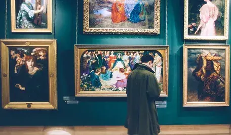 Mężczyzna w zielonym płaszczu ogląda obrazy wiszące na ścianie w eleganckiej galerii sztuki, otoczonej bogato zdobionymi, złotymi ramami.