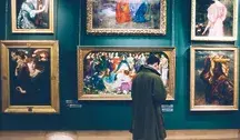 Mężczyzna w zielonym płaszczu ogląda obrazy wiszące na ścianie w eleganckiej galerii sztuki, otoczonej bogato zdobionymi, złotymi ramami.