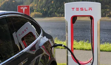 Samochód elektryczny Tesla podłączony do ładowarki Supercharger, widoczna stacja ładowania z logo Tesli na tle jeziora i lasu.