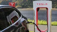 Samochód elektryczny Tesla podłączony do ładowarki Supercharger, widoczna stacja ładowania z logo Tesli na tle jeziora i lasu.