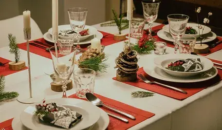 Świątecznie nakryty stół wigilijny z białą zastawą, czerwonymi bieżnikami, świecami, szyszkami i naturalnymi dekoracjami bożonarodzeniowymi.