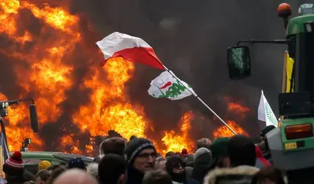 Rolnicy protestujący w Brukseli. W centralnym punkcie polska flaga, na tle płomieni ognia, obok widoczny jest ciągnik rolniczy.